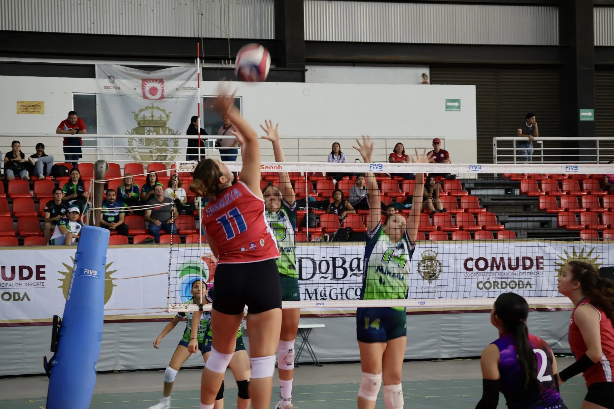 Una jugadora de rojo ataca en la red contra dos rivales de verde.