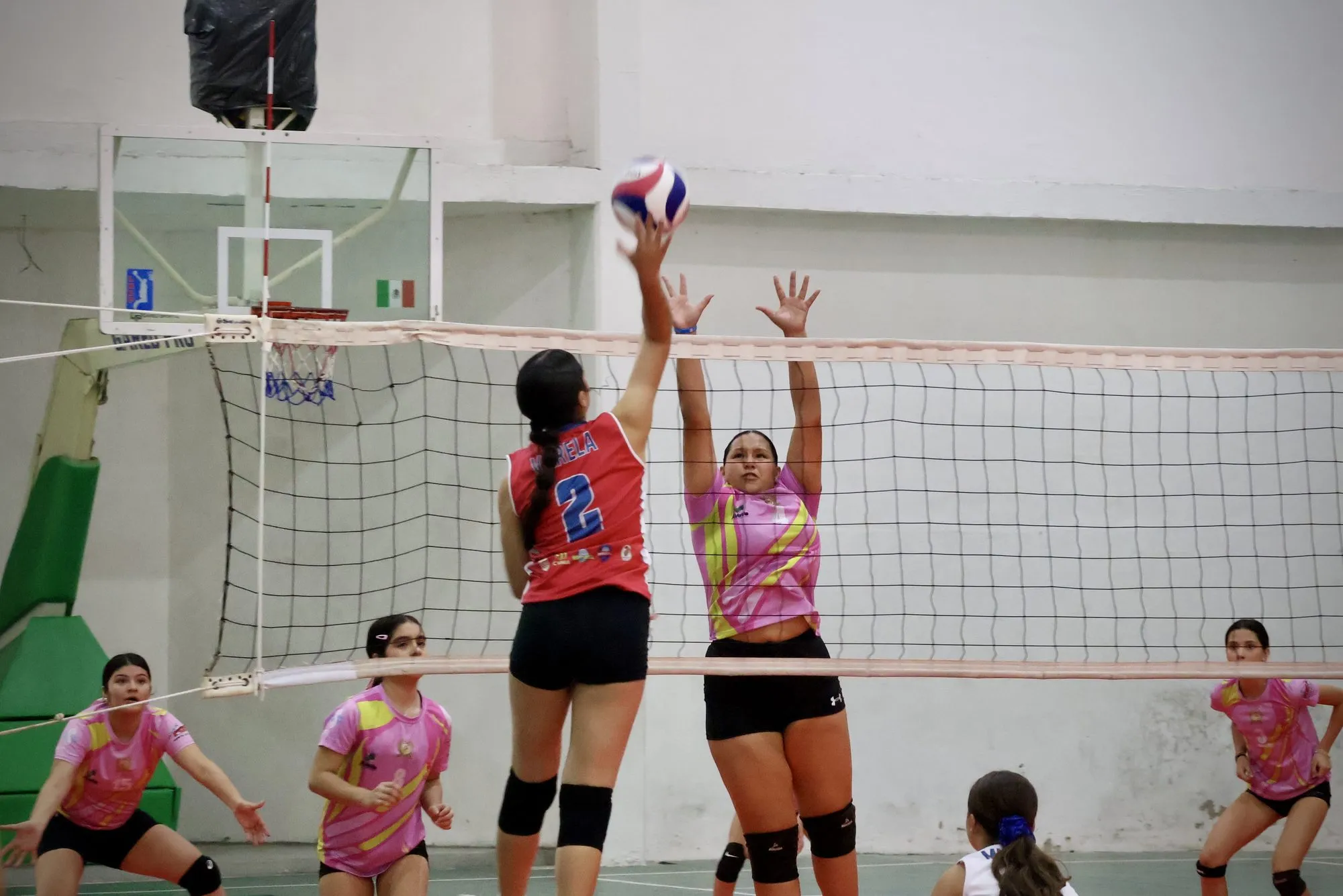 Dos jugadoras se enfrentan en la red, una de rojo y otra de rosado, con el balón entre ellas.