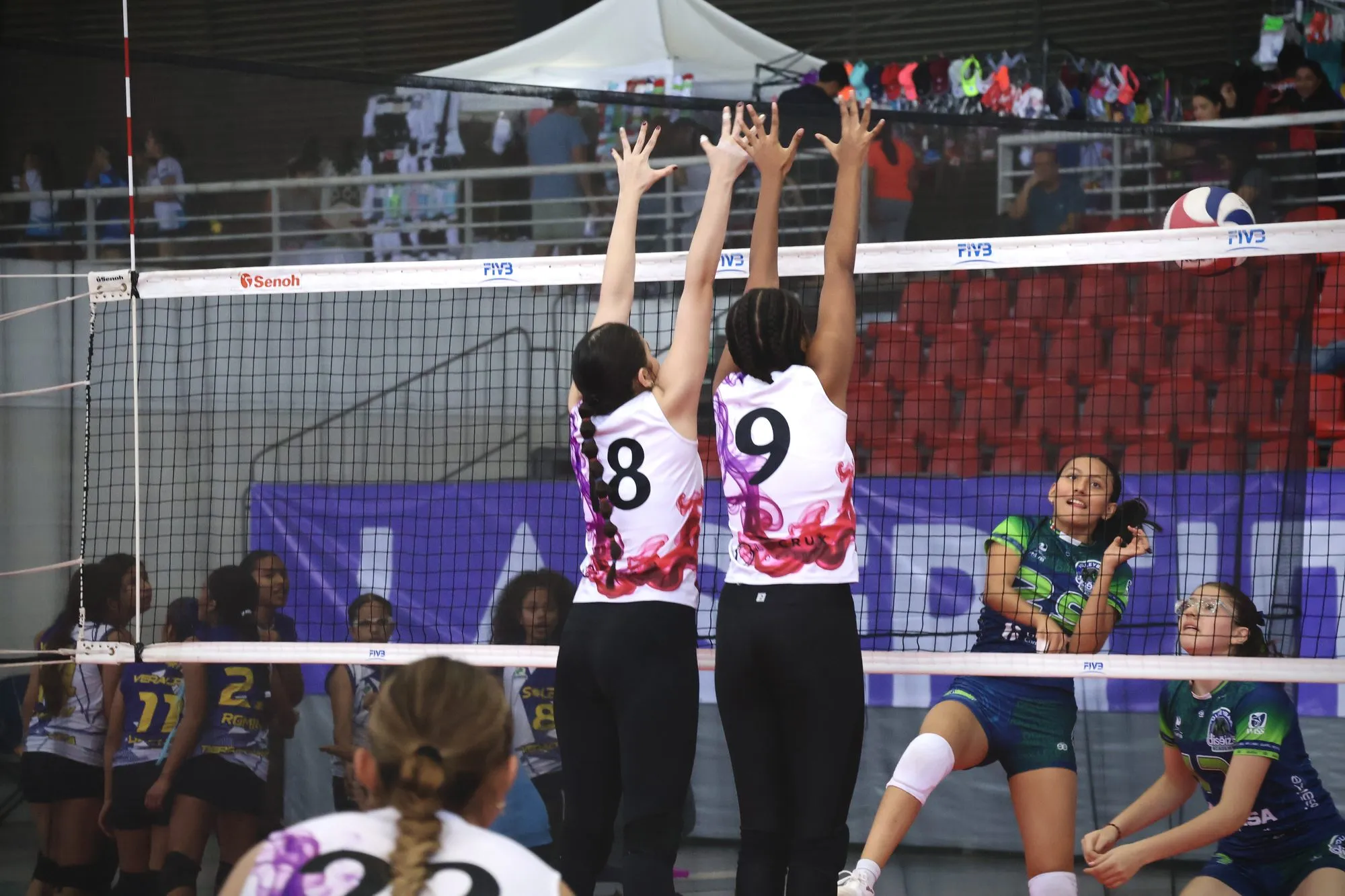 Dos bloqueadoras de blanco levantan las manos en la red ante el equipo verde.
