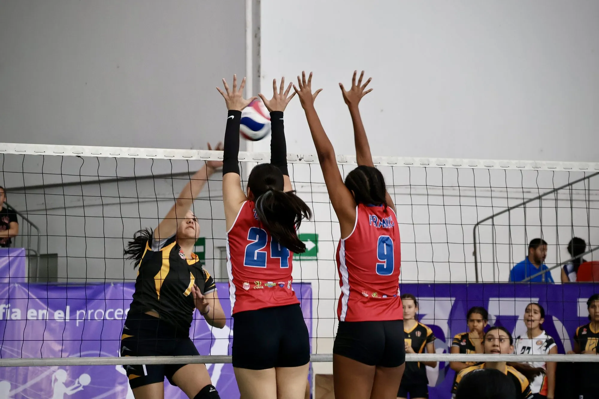 Dos jugadoras de rojo saltan juntas a bloquear un remate en la red.