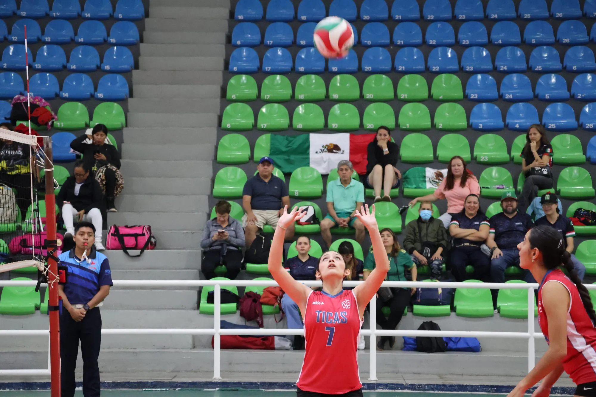Una colocadora levanta el balón con los dedos mientras la gradería verde y azul observa.