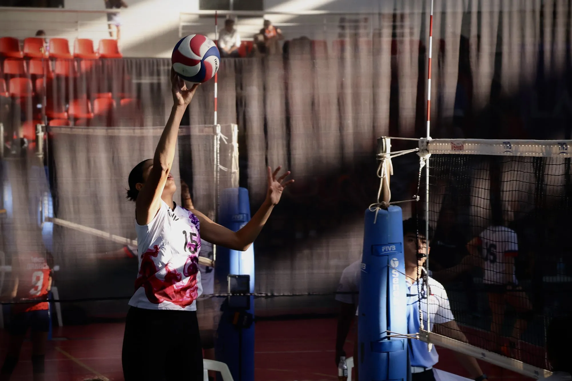 Una jugadora coloca el balón junto al poste de la red, con el gimnasio en penumbra.