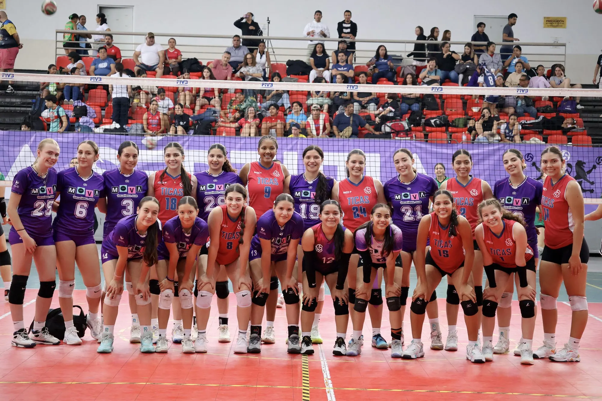 Los dos equipos posan juntos a un lado de la red al terminar el partido, uno de morado y otro de rojo.