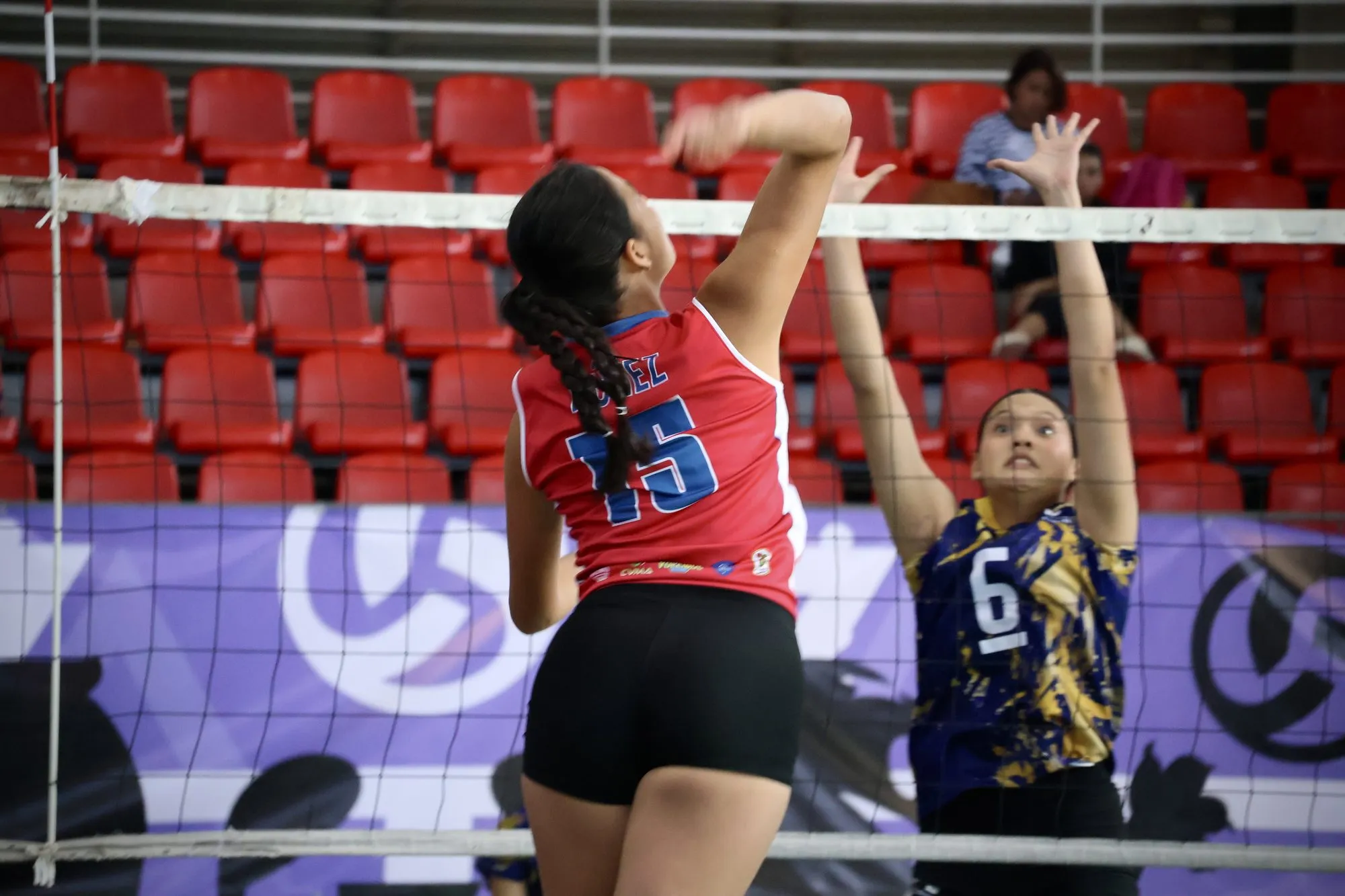 Una atacante de rojo remata ante una bloqueadora del equipo azul y dorado.