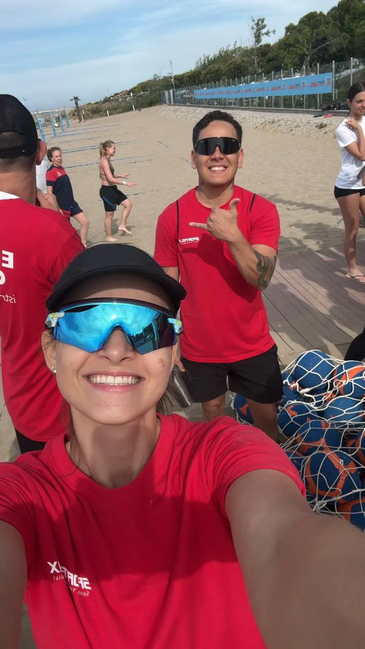 Una entrenadora sonríe a cámara en la arena, con el resto del grupo trabajando detrás.